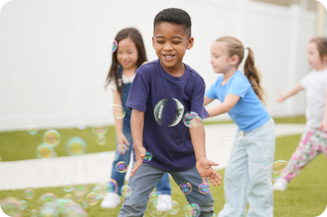 A boy playing with bubbles outside with other children in the background.