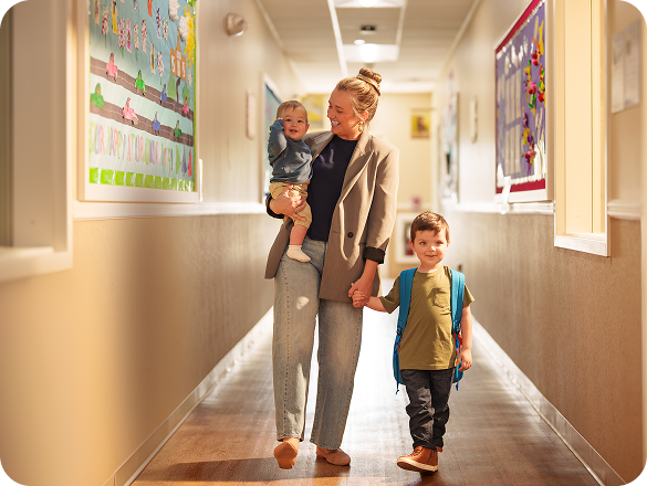A mother holding a baby and holding the hand of an older child while walking down a hallway.