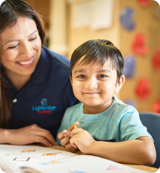 A preschooler smiling at the camera while a teacher looks on.