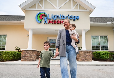 A mom holding a baby and the hand of a young boy walking out of a Lightbridge Academy.