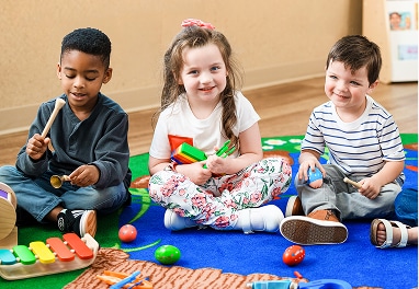 Three children sitting on the floor in a classroom playing musical instruments.