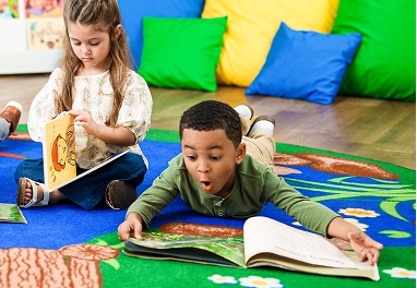 A boy reading a book while lying on the floor in a classroom.