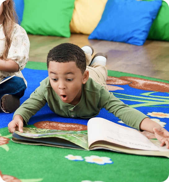 A boy laying on the floor reading a book with a surprised expression on his face.