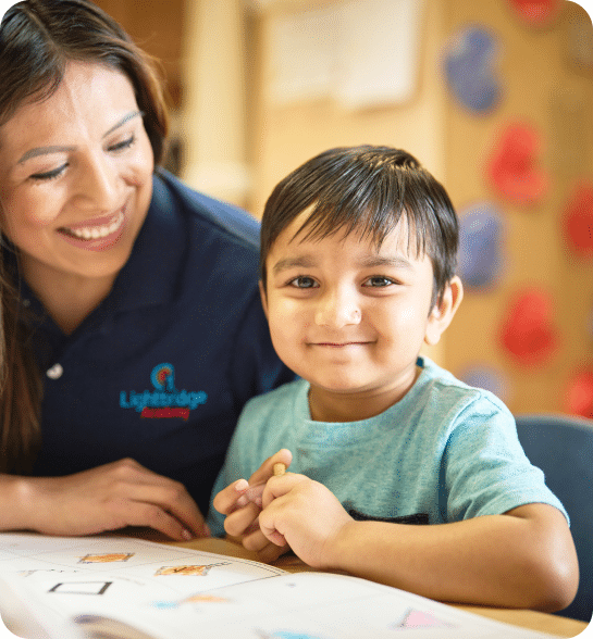 A preschooler smiling at the camera while a teacher looks on.