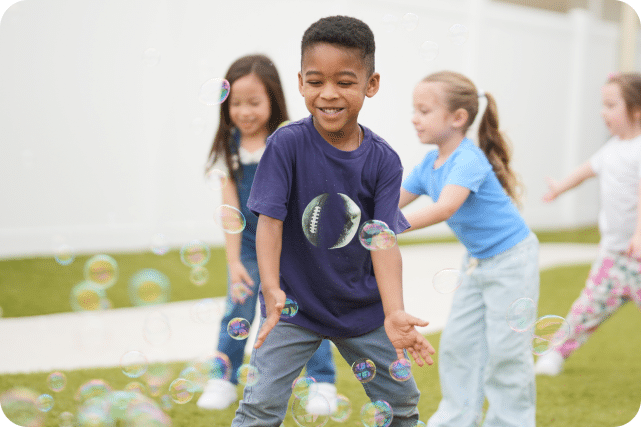 A boy playing with bubbles outside with other children in the background.