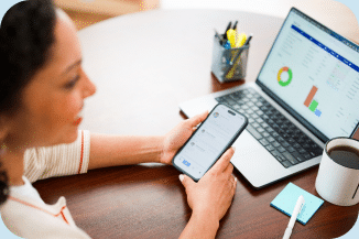 A woman looking at a smart phone while working on a laptop.