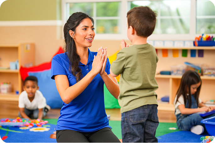 A teacher kneels on the floor facing a young child while clapping her hands.