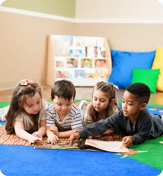 Four children reading a book while lying on a rug in a classroom.