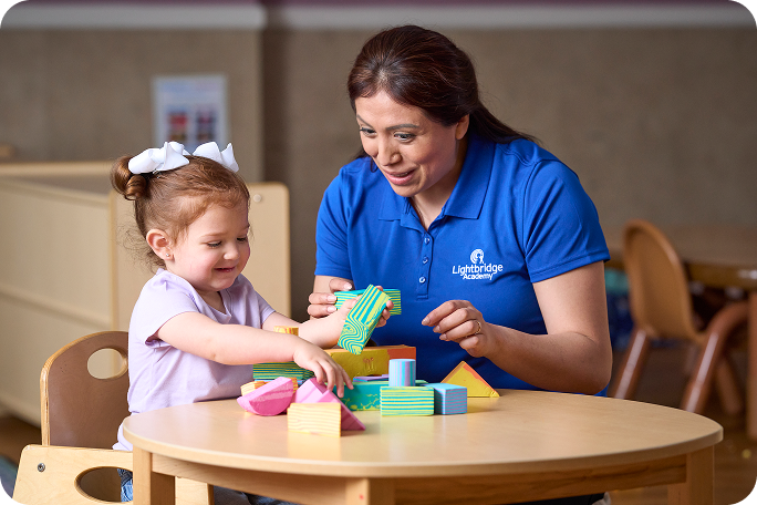 A teacher playing blocks with a young girl while seated at a table.